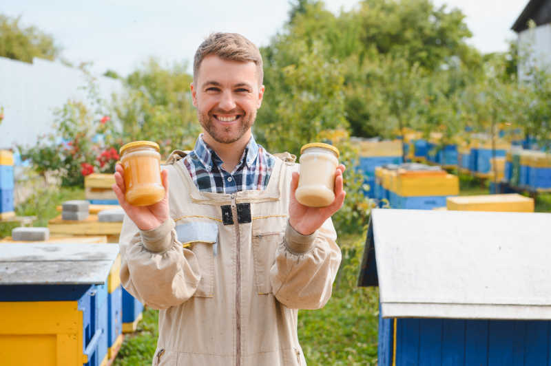 A beekeeper holding two jars of honey with beehives in the field behind him