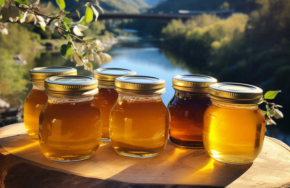 Jars of extracted liquid honey on a bench outside with a river in the background