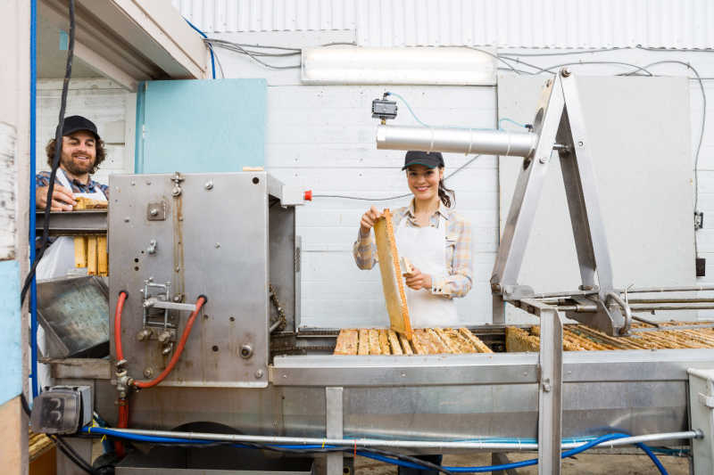 Two apiary workers smiling while they work next to a large commercial honey extraction machine